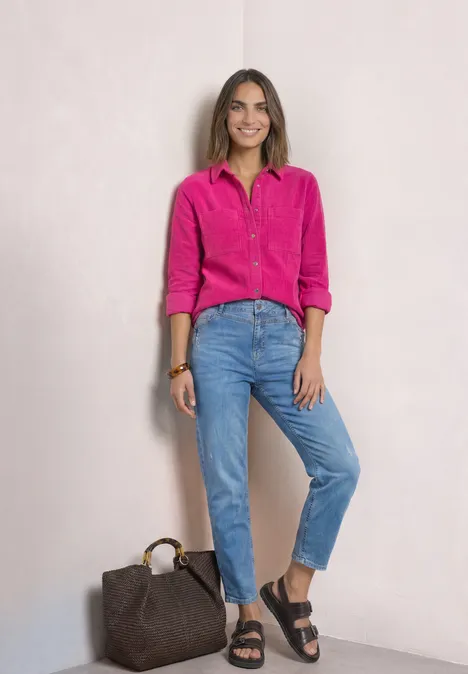Corduroy overhemd met lange mouwen en knopen magenta dream Frau in Jeans und pinker Bluse lehnt vor heller Wand, neben ihr eine braune Tasche.