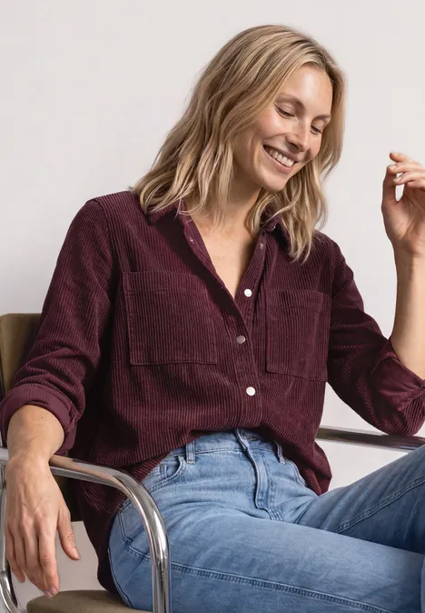 Corduroy overhemd met lange mouwen en knopen rustic rouge Eine lächelnde Frau in weinrotem Cord-Hemd und Jeans sitzt entspannt auf einem Stuhl.