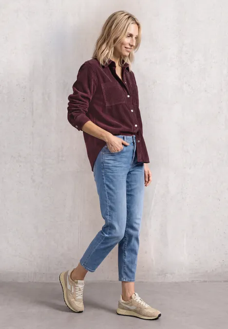 Corduroy overhemd met lange mouwen en knopen rustic rouge Frau im bordeauxfarbenen Cordhemd, hellblauen Jeans und beigen Sneakern vor hellem Hintergrund.