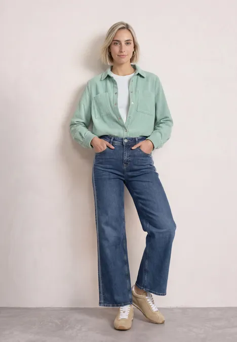Corduroy overhemd met lange mouwen en knopen eucalyptus green Frau vor neutralem Hintergrund in mintgrünem Hemd, weißem Top, weitem Jeans und Sneakern.