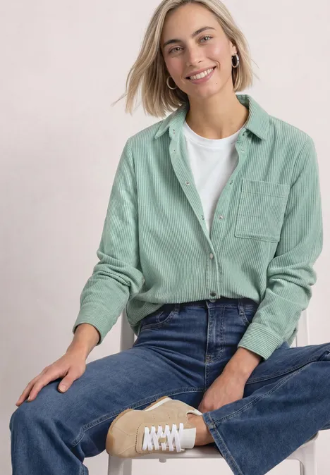 Corduroy overhemd met lange mouwen en knopen eucalyptus green Frau sitzt im Schneidersitz, lächelt, trägt mintgrünes Hemd, weißes T-Shirt, Jeans und beige Sneaker.
