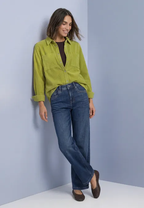 Corduroy overhemd met lange mouwen en knopen apple lime Frau lehnt an Wand, trägt grüne Bluse, dunkles Top, blaue Jeans und braune Schuhe.