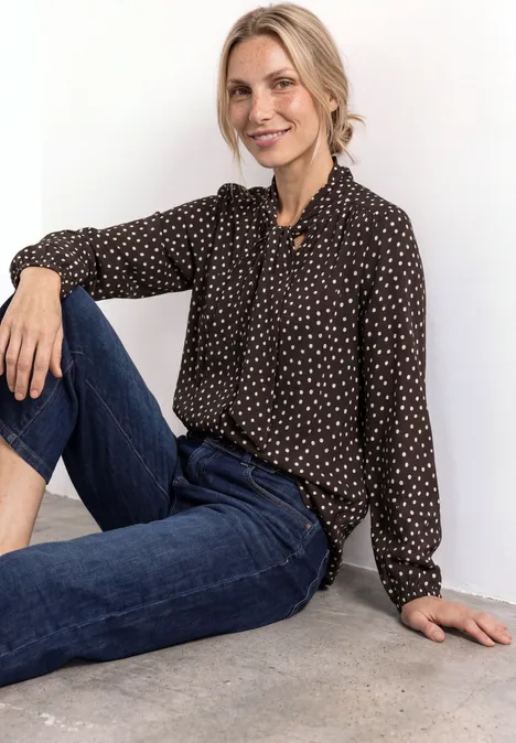Langarm Bluse mit Schleifendetail black coffee Frau sitzt lässig am Boden, trägt schwarze gepunktete Bluse und Jeans, lehnt an weißer Wand.