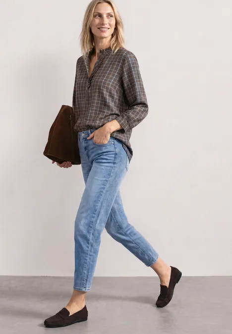 Langarm Bluse mit Rüschendetails silk brown Eine lächelnde Frau in kariertem Hemd, Jeans und Mokassins hält eine braune Tasche.