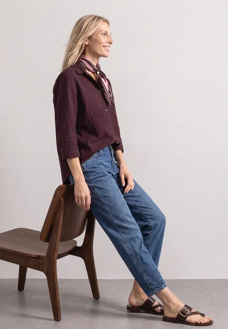 Overhemd met 3/4-mouwen en corduroy-mix rustic rouge Een vrouw leunt tegen een stoel, draagt spijkerbroek, bordeaux overhemd, zwarte sandalen en lacht opzij.