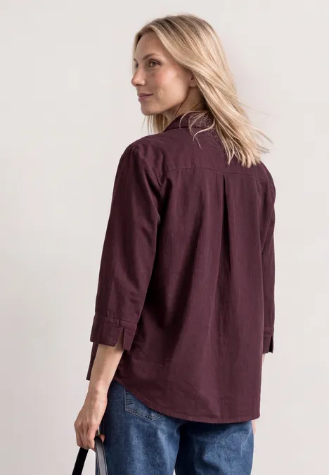 Overhemd met 3/4-mouwen en corduroy-mix rustic rouge Vrouw van opzij kijkt over haar schouder, draagt een bordeaux overhemd en blauwe spijkerbroek.