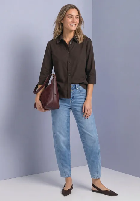 Overhemd met 3/4-mouwen en corduroy-mix black coffee Vrouw in een bruine blouse, lichtblauwe jeans, zwarte pumps en een bordeauxrode tas tegen een grijze muur.