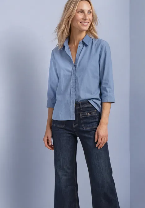 Vrouw lacht in lichtblouse en donkere jeans voor een lichte achtergrond.