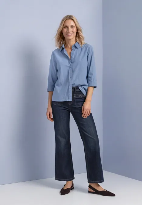Vrouw voor neutrale achtergrond, draagt lichtblauw overhemd, wijde enkellange jeans en platte schoenen.