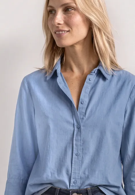 Een blonde vrouw draagt een lichtblauw overhemd met lange mouwen voor een lichte achtergrond.