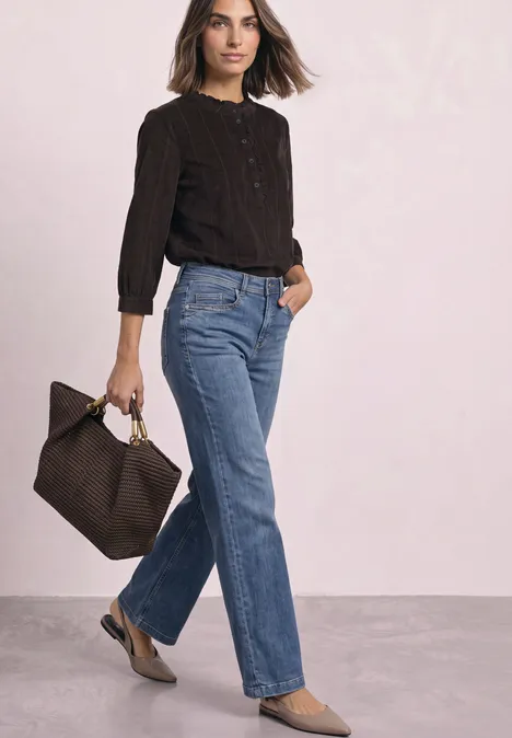 Eine Frau in dunklem Pullover und geraden Jeans, hält eine Tasche in der Hand.