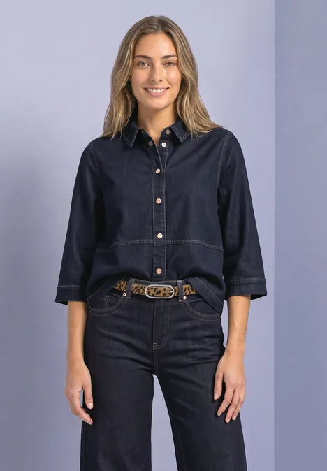 Frau in dunklem Jeans-Outfit mit 3/4-Ärmeln und Ziergürtel, lächelnd und frontal stehend.