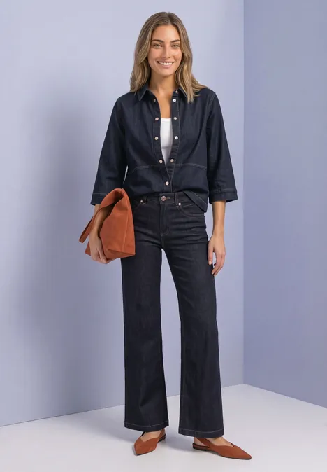 Frau in dunklem Denim-Hemd und Schlaghose mit weißem Top, brauner Tasche und spitzen Schuhen vor hellem Hintergrund.
