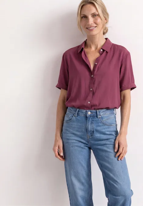 Overhemd met korte mouwen in effen kleur dark clouded blush Frau steht vor weißer Wand, trägt brombeerfarbene Bluse und lockere Jeans, lächelt.