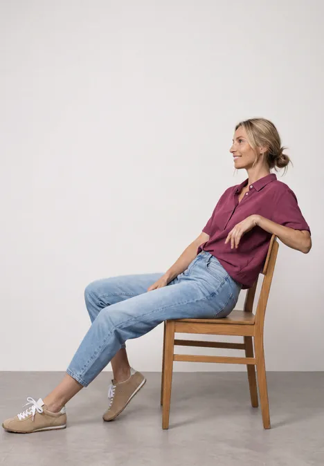 Overhemd met korte mouwen in effen kleur dark clouded blush Frau sitzt seitlich auf Holzstuhl, lässig zurückgelehnt, trägt violettes Shirt, Jeans und Sneaker.