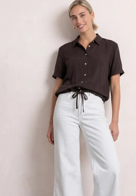 Overhemd met korte mouwen in effen kleur black coffee Frau in lockerer brauner Bluse und weißen, weiten Bundfaltenhosen mit Taillenkordel.