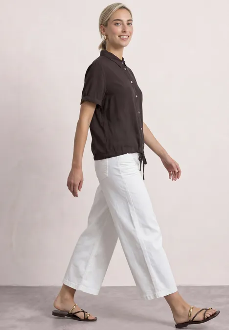 Overhemd met korte mouwen in effen kleur black coffee Eine Frau in schwarzem Oberteil und weißen Hosen geht lächelnd in Sandalen.