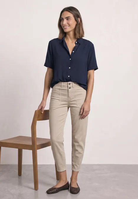 Overhemd met korte mouwen in effen kleur darkest blue Frau in marineblauem Kurzarmhemd, beigen Hosen und Loafers, stehend neben einem Stuhl vor hellem Hintergrund.