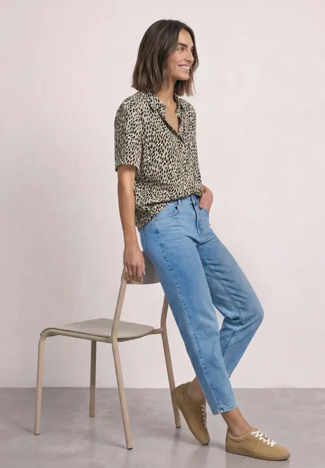 Kurzarm Hemdbluse mit Print cotton beige Frau in Leopardenmuster-Bluse, blauen Jeans und beigen Schuhen lehnt lässig an einem Stuhl.