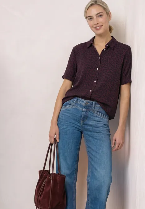 Eine lächelnde Frau trägt ein gepunktetes Hemd, Jeans und hält eine rote Wildledertasche vor hellem Hintergrund.
