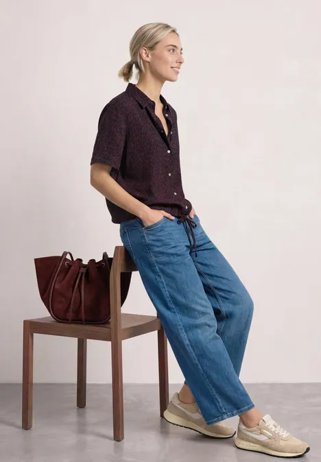 Eine Frau lehnt an einem Stuhl, trägt weite Jeans, kurzärmliges Hemd, Sneakers, neben ihr eine Handtasche.