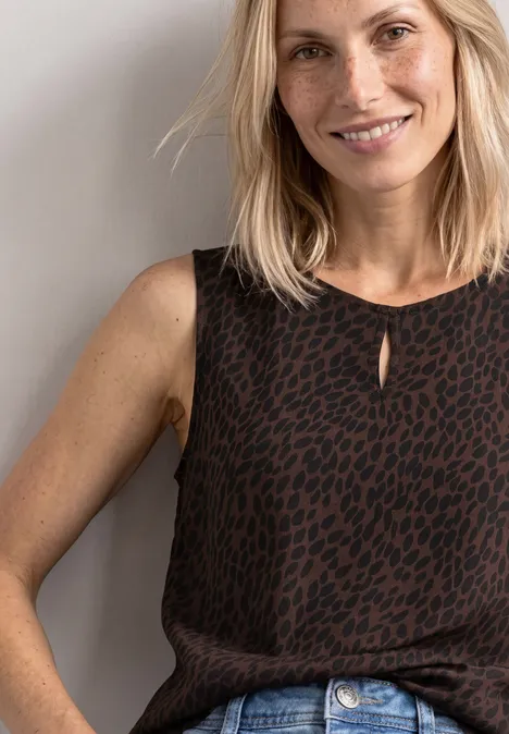 Frau mit blondem Haar und braunem, gemustertem ärmellosem Oberteil, lächelnd vor hellem Hintergrund.