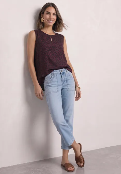 Jonge vrouw leunt tegen een muur, draagt een mouwloze top met stippen, lichte jeans en bruine sandalen.