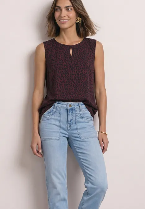 Vrouw lacht, draagt een mouwloze rood-zwarte print top en lichtblauwe jeans, staande voor een lichte achtergrond.