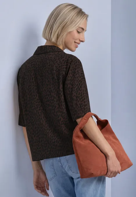 Frau mit kurzem blondem Haar in braun gemusterter Bluse und Jeans, hält orange-braune Tasche seitlich.