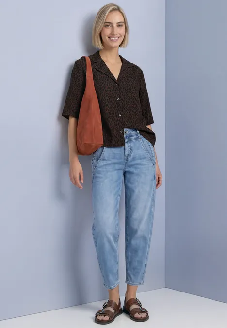Frau mit kurzem blondem Haar trägt schwarz gepunktete Bluse, helle Jeans, braune Sandalen und eine orange Tasche.