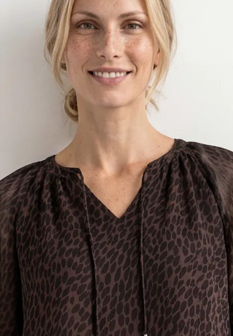 Eine lächelnde Frau in brauner, gemusterter Bluse vor hellem Hintergrund.