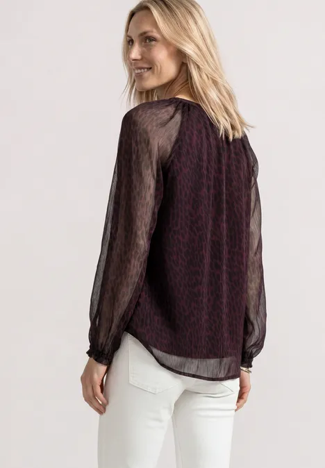 Eine Frau trägt eine dunkelrote Bluse mit transparenten schwarzen Ärmeln und weiße Hose.