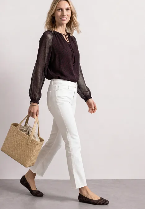 Frau in weißer Hose, schwarzer Bluse, trägt beige Tasche und flache Loafers.