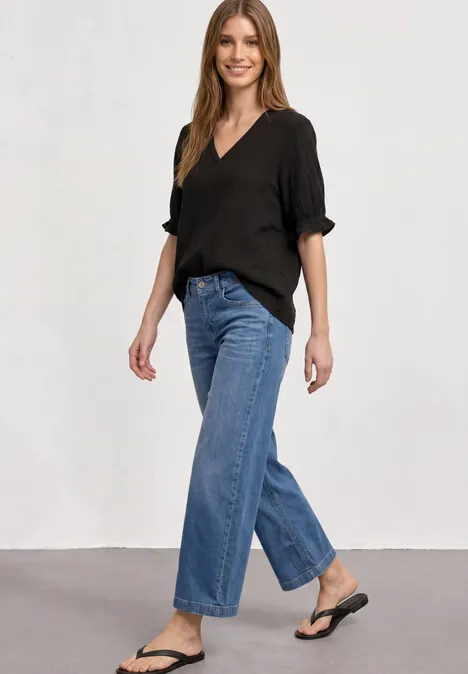 Frau in schwarzem V-Neck-Oberteil, weiten Jeans und schwarzen Flip-Flops vor schlichtem Hintergrund.