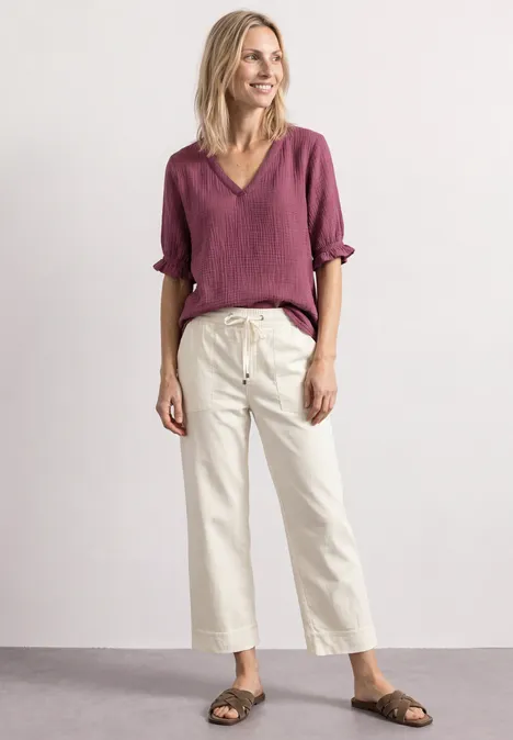 Eine lächelnde Frau in roter Bluse, weißer Culotte und Sandalen vor neutralem Hintergrund.