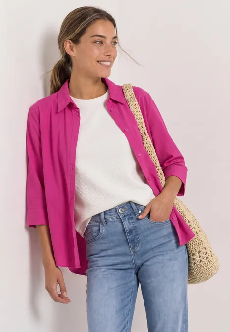 Eine lächelnde junge Frau in pinkem Hemd, weißem T-Shirt und Jeans, mit einer Strohtasche.