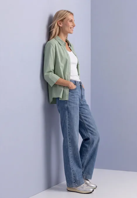 Frau lehnt lässig an Wand, trägt grünes Sakko, weißes Shirt, weite Jeans und silberne Sneaker.