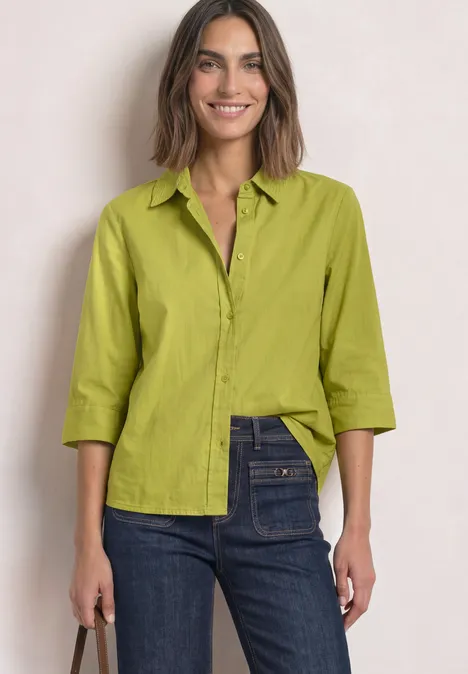 Eine Frau mit einem hellgrünen Hemd und blauen Jeans lächelt leicht.