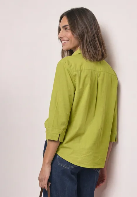 Frau mit schulterlangem braunem Haar in grüner Bluse und Jeans, seitlich stehend und lächelnd.