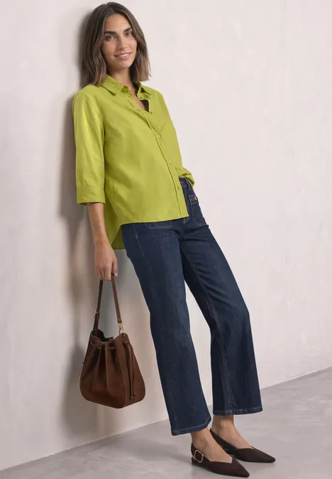 Frau in grüner Bluse und Jeans, braune Tasche, lässig an Wand gelehnt.