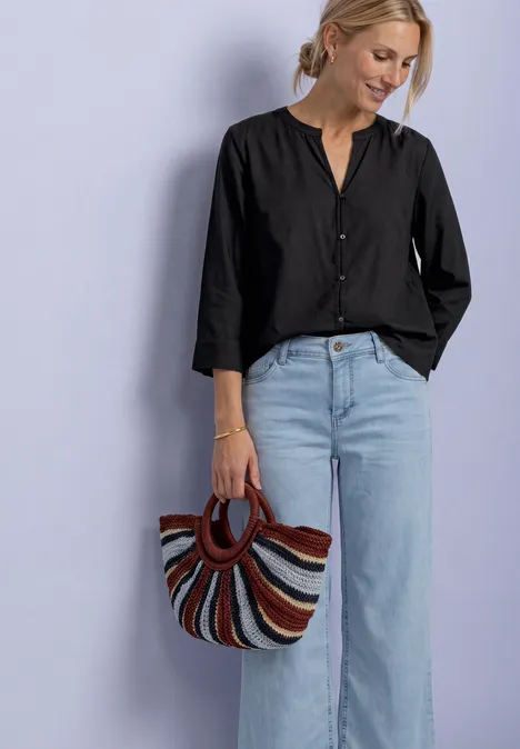 Blouse met 3/4-mouwen en split in de hals Black Vrouw in zwart bloesje en lichtblauwe jeans met gekleurde gestreepte tas voor paarse achtergrond.