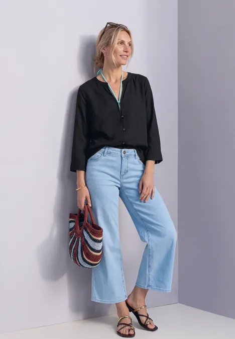 Blouse met 3/4-mouwen en split in de hals Black Vrouw leunt tegen muur, zwarte blouse, wijde lichte jeans, zwarte sandalen en kleurrijke gestreepte tas.