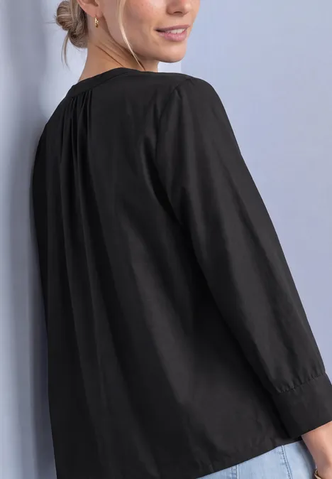 Blouse met 3/4-mouwen en split in de hals Black Vrouw met blond haar in een staart draagt een losse zwarte blouse met lange mouwen en lacht.