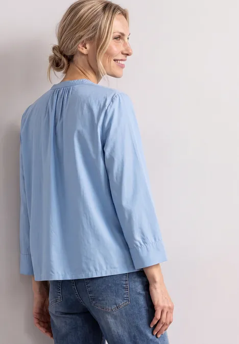 Femme souriante en chemise bleue claire et jean, vue de côté sur fond lumineux.