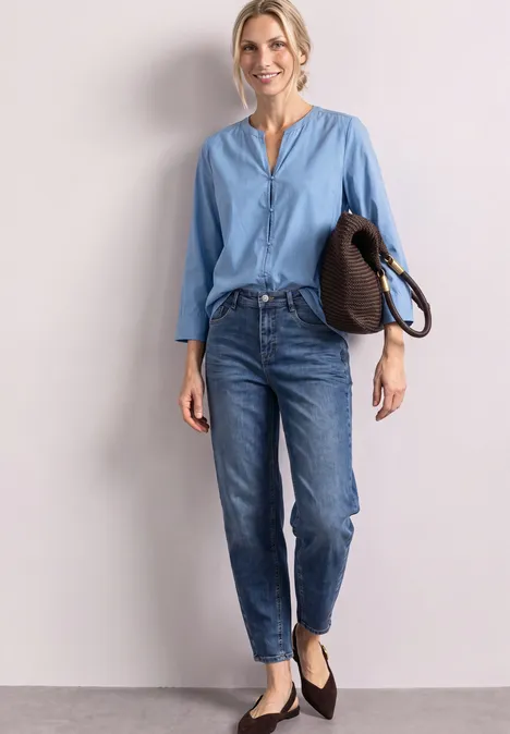 Jeune femme devant un mur clair, portant une blouse bleue, un jean et des ballerines noires, tenant un sac.