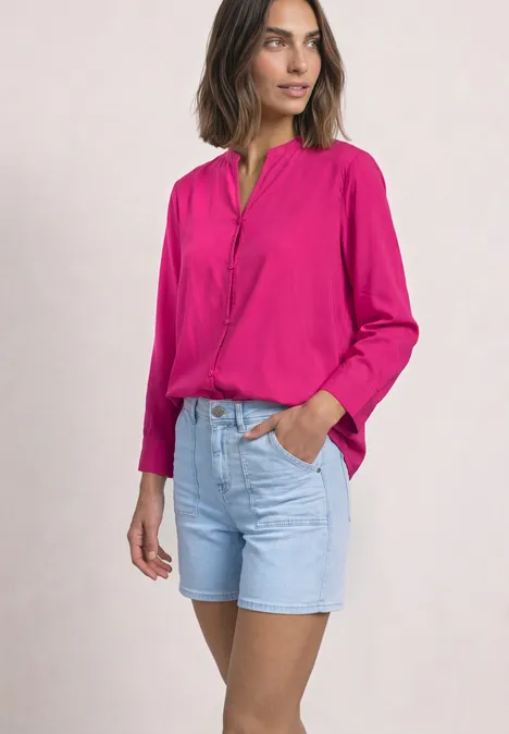 Frau trägt pinke Bluse und hellblaue Jeans-Shorts, steht lässig mit einer Hand in der Tasche.