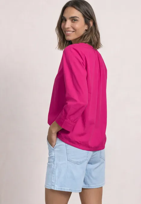 Frau in pinkem Langarmshirt und hellblauen Jeans-Shorts blickt lächelnd über die Schulter.