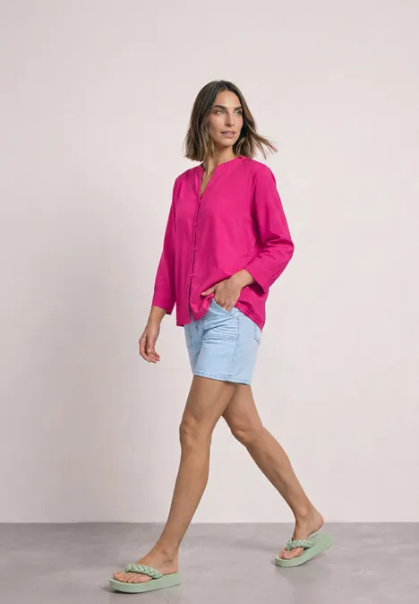 Eine Frau in pinker Bluse, hellblauen Jeans-Shorts und mintgrünen Sandalen geht seitlich vor hellem Hintergrund.