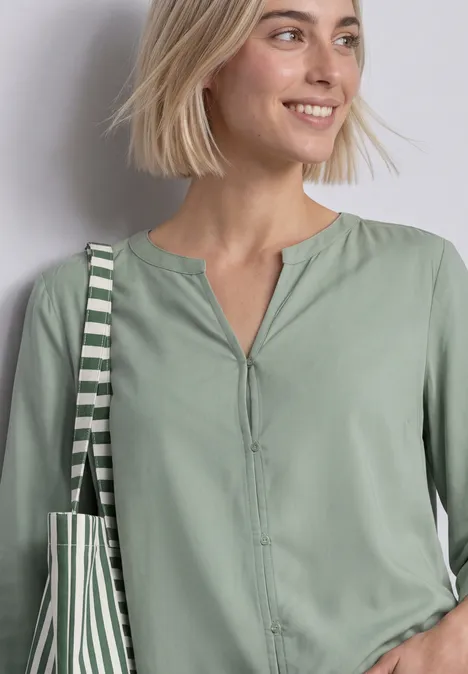 Blouse met 3/4-mouwen en split in de hals eucalyptus green Vrouw met kort blond haar draagt lichtgroen overhemd en gestreepte tas, glimlachend.