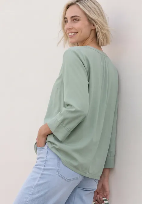 Blouse met 3/4-mouwen en split in de hals eucalyptus green Glimlachende blonde vrouw in lichtgroene longsleeve en blauwe jeans, staand opzij voor een lichte muur.